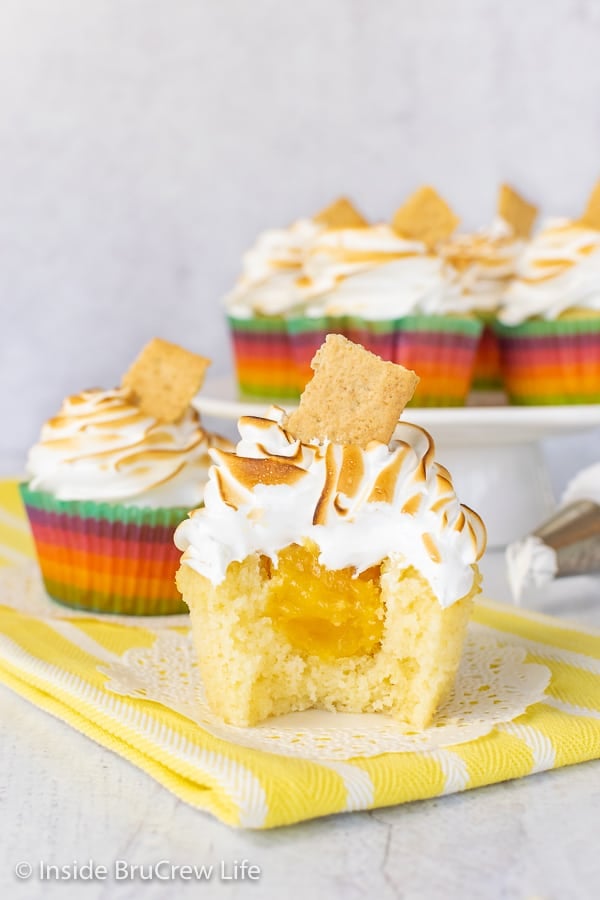 Two lemon meringue cupcakes on white doilies with a bite out of the front cupcake showing the hidden lemon curd center and a cake plate with more cupcakes behind them