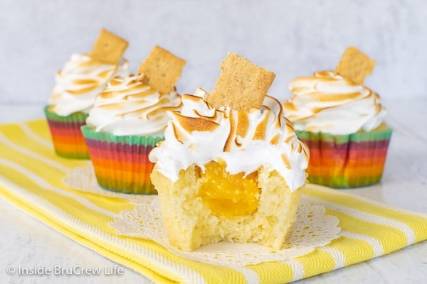 Four lemon meringue cupcakes on a yellow towel with a bite taken out of the front vanilla cupcake showing the hidden lemon curd center