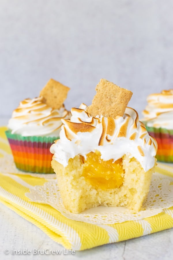Three Lemon Meringue Cupcakes sitting on white doilies on a yellow towel with a bite taken out of the front one showing the hidden lemon center.