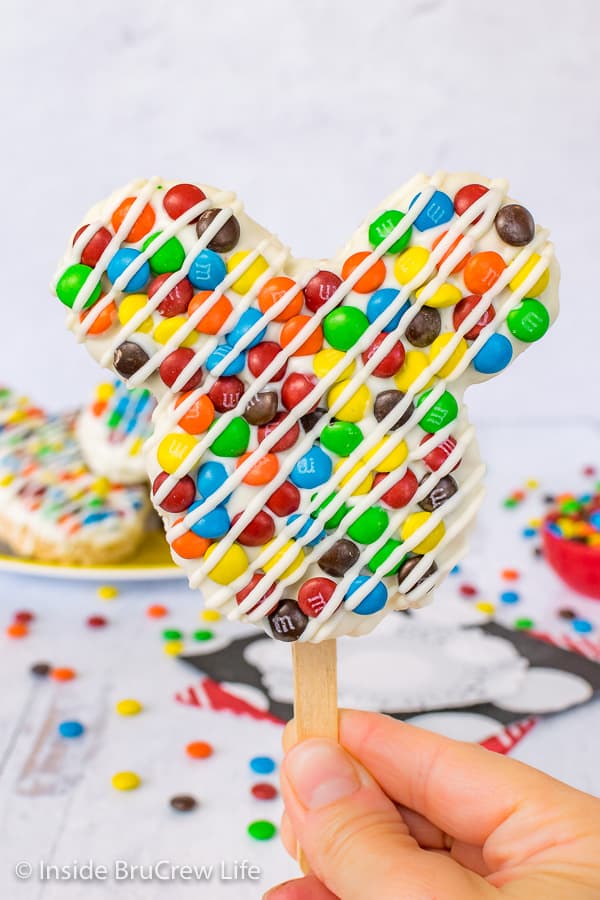 A close up picture of a M&M's Mickey Mouse Rice Krispie Treat on a stick held up in front of a plate of treats