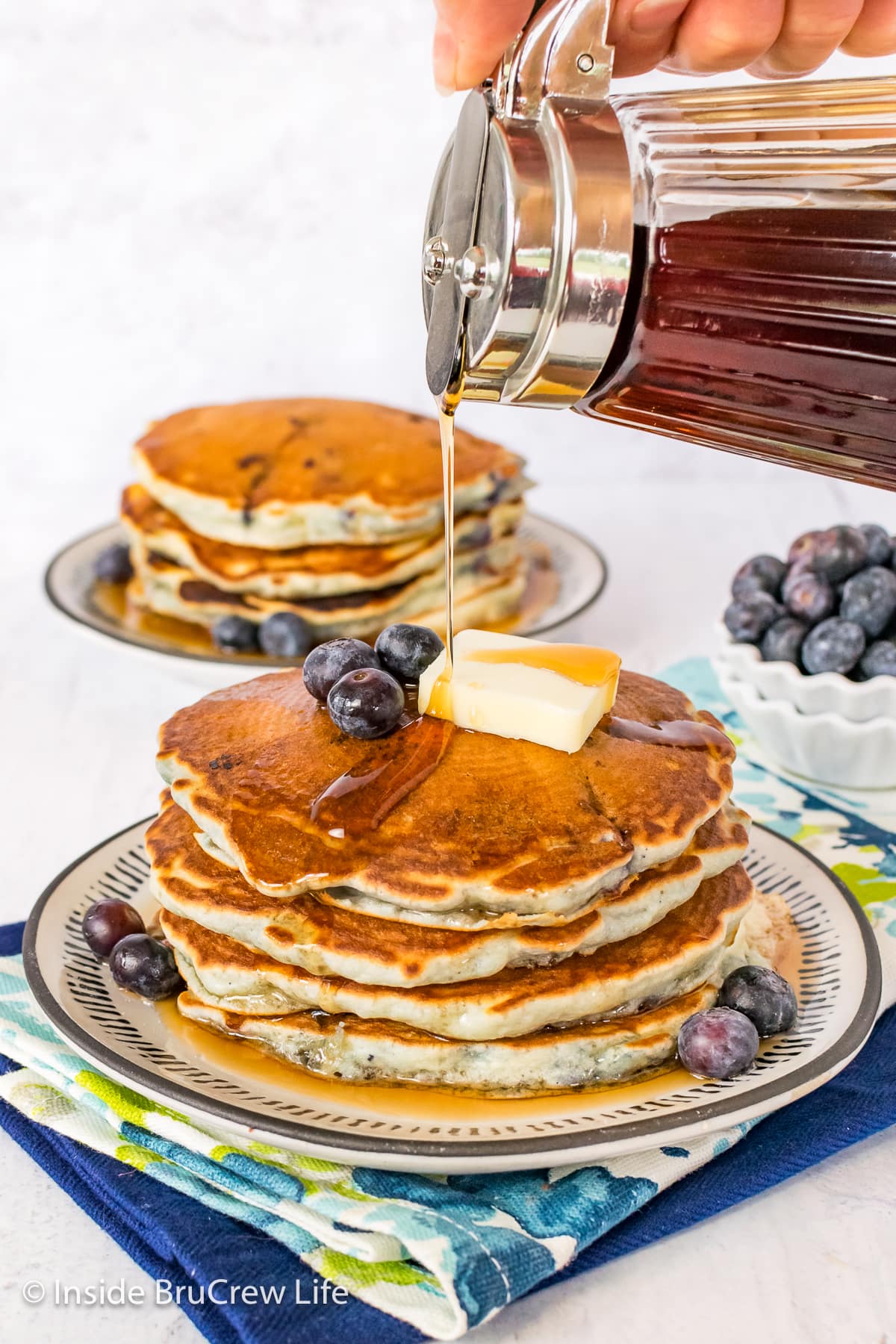 A stack of blueberry pancakes on a plate with syrup being drizzled on top.