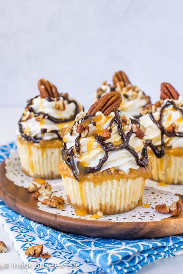 A brown wooden plate with four mini turtle cheesecakes topped with whipped cream and chocolate and caramel drizzles