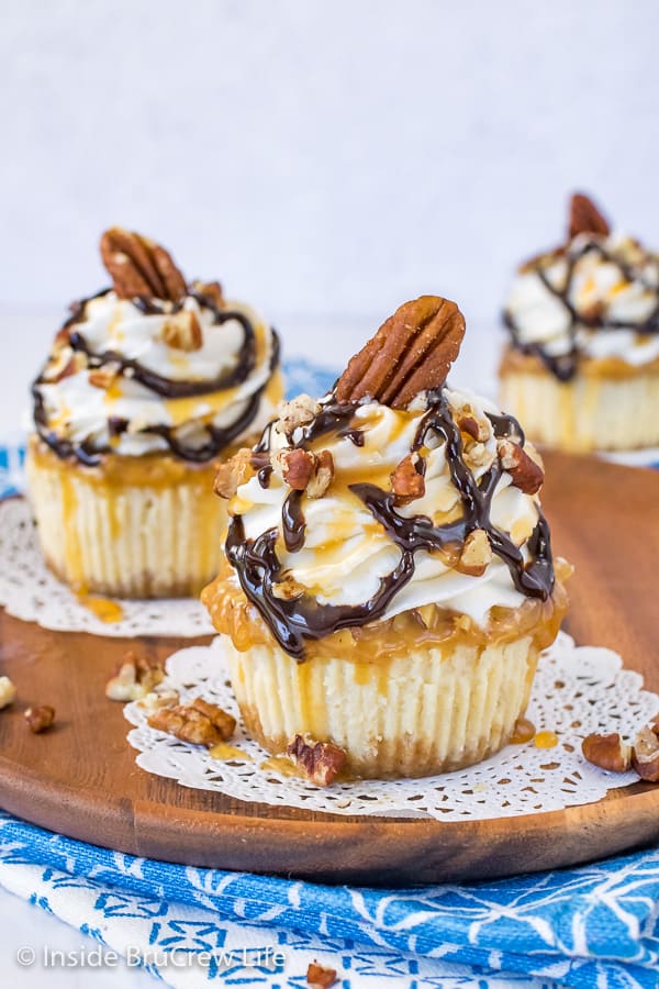 Two mini turtle cheesecakes topped with whipped cream and caramel and chocolate drizzles on a brown plate