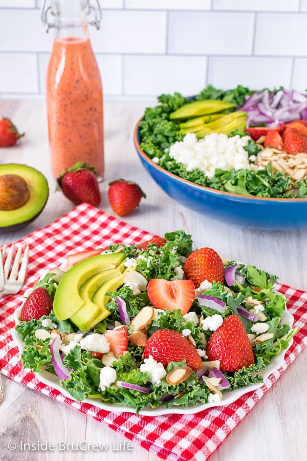 A white plate filled with a strawberry avocado kale salad and a big bowl of salad behind it