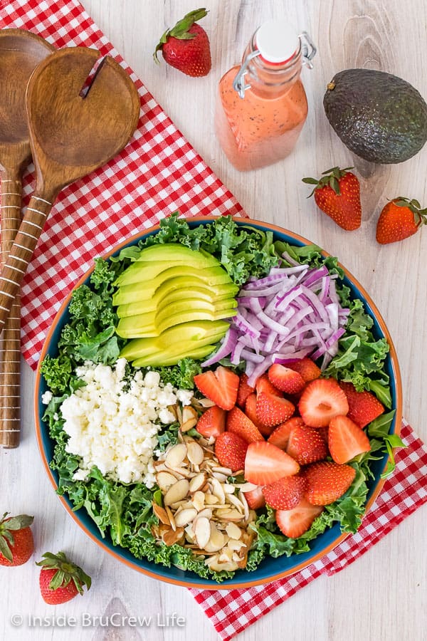 A large salad bowl on a red and white towel filled with kale and topped with avocados, onions, strawberries, nuts, and cheese