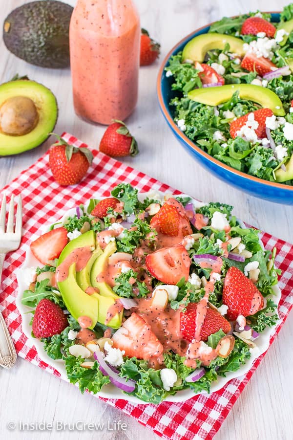 A white plate on a red and white checkered towel filled with strawberry avocado kale salad and drizzled with strawberry poppy seed dressing