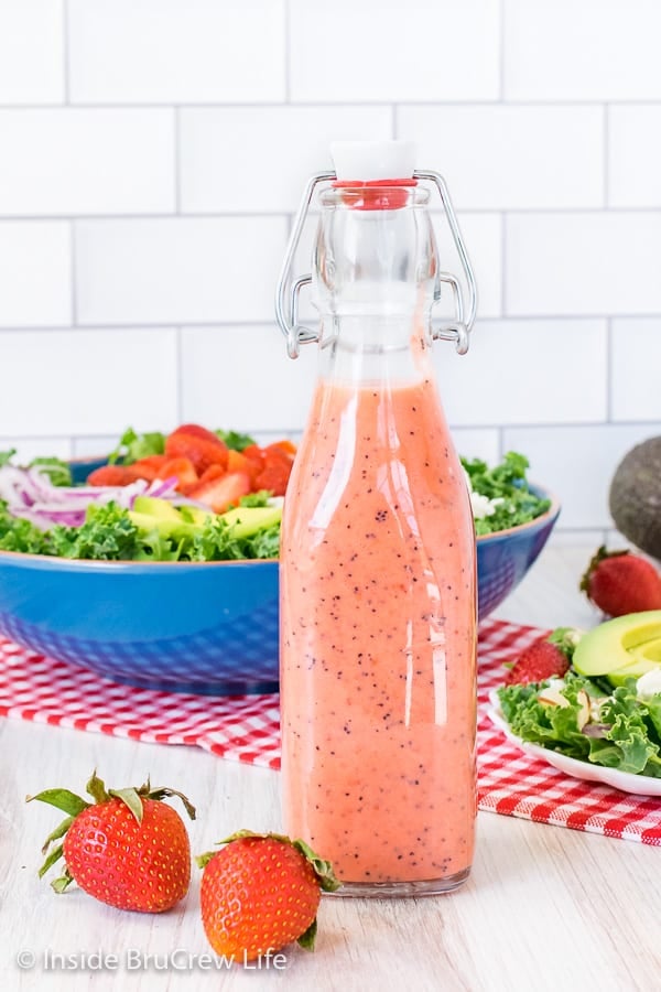 A full bottle of strawberry poppy seed salad dressing with a bowl of strawberry kale salad behind it