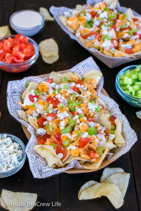 A close up of a cardboard container filled with buffalo chicken chips and bowls of toppings around it