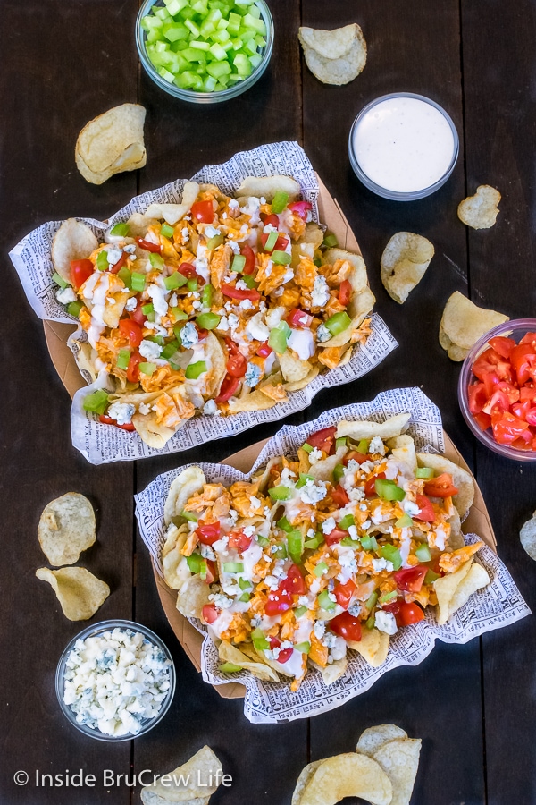 Two cardboard containers on a dark background filled with buffalo chicken chips that are topped with ranch dressing and bleu cheese crumbles