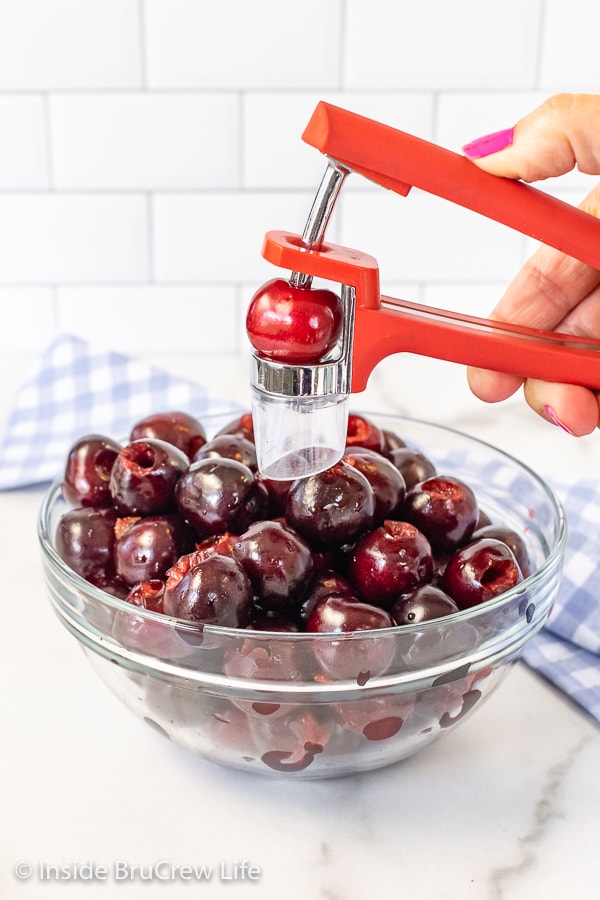 A bowl of fresh cherries with a cherry pitter held over the bowl with a cherry in it