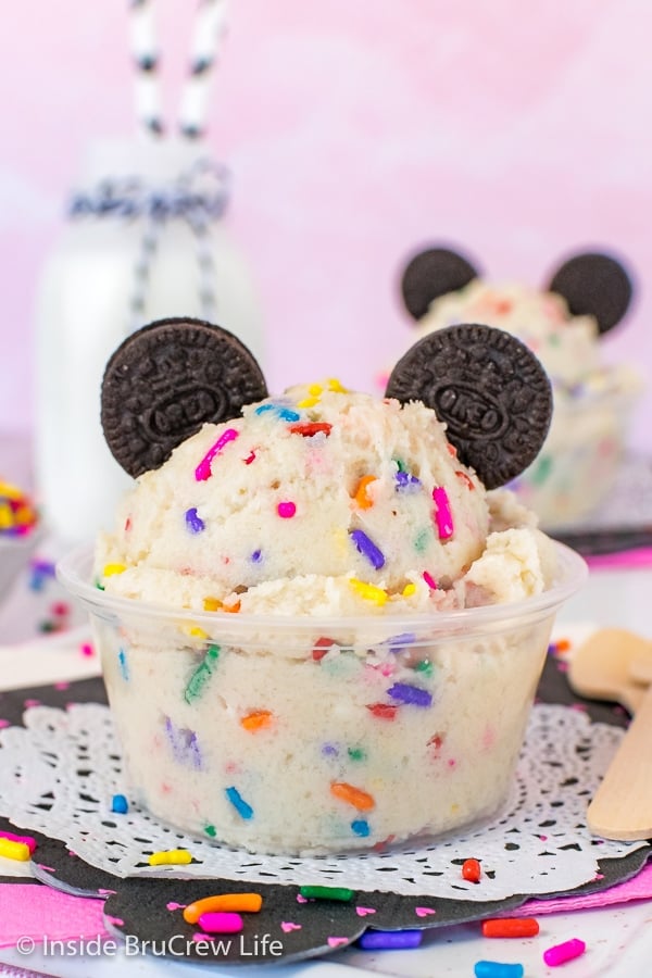 A close up picture of a clear cup filled with cookie dough with sprinkles and chocolate cookie Mickey Mouse ears.