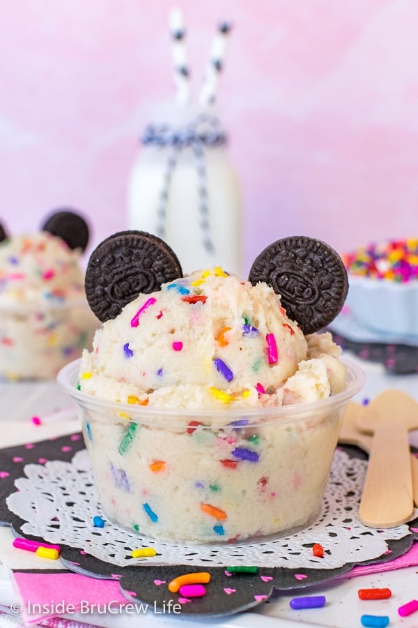 A clear cup with cookie dough made with rainbow sprinkles and topped with chocolate cookie Mickey Mouse ears.