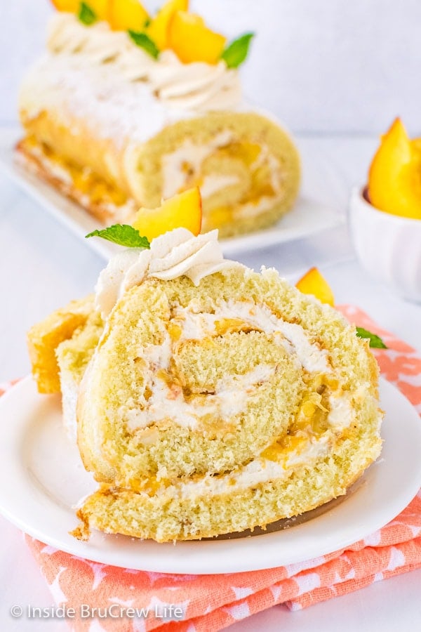 A white plate with a slice of peaches and cream cake roll on it and the rest of the cake roll behind it