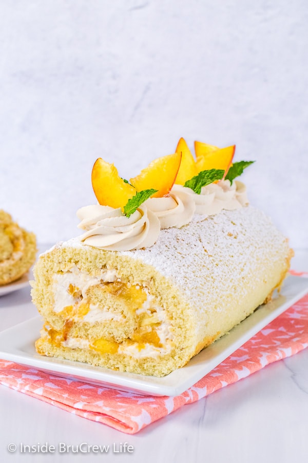 A white serving plate with a Peaches and Cream Cake Roll on it decorated with whipped cream swirls and fresh peach slices