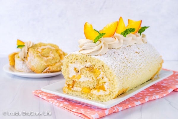 A white plate with a peaches and cream cake roll on it decorated with whipped cream swirls and fresh peach slices