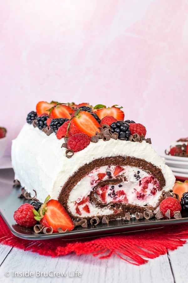 A full Berries and Cream Chocolate Cake Roll on a black plate with the end showing the swirl of cake, whipped cream, and berries