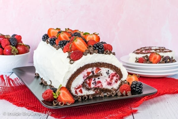 A black plate with berries and cream chocolate cake roll on it showing the inside and a slice on a plate behind it