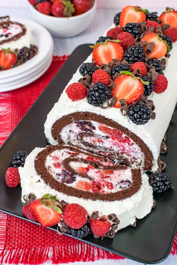 Overhead picture of a berries and cream chocolate cake roll on a black plate with one slice flipped down so you see the cream and berry filling.