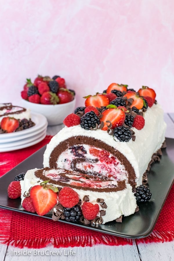 A black plate on a red towel with a berries and cream chocolate cake roll on it and one slice lying down at the front