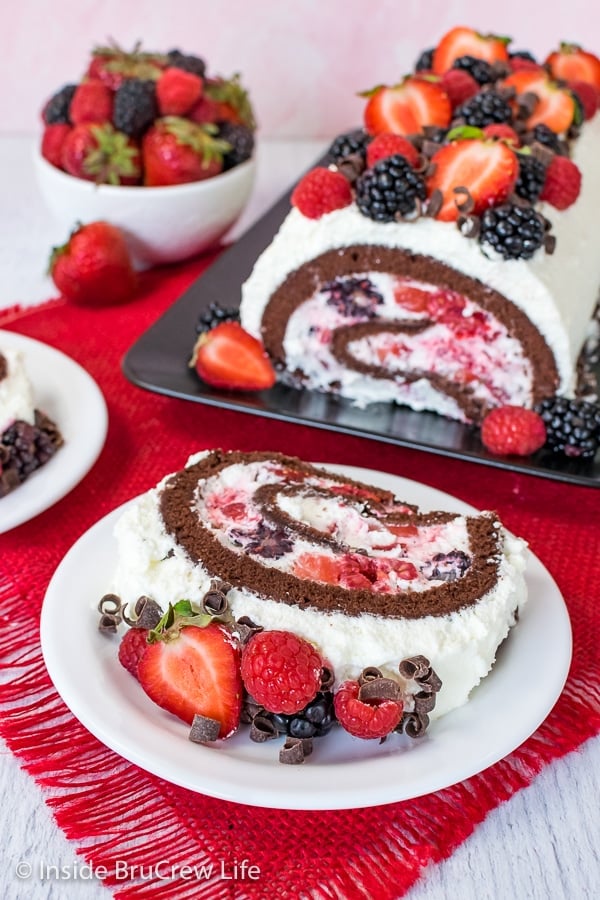 A white plate with a slice of berries and cream chocolate cake roll on it and the rest of the cake roll behind it on a black plate