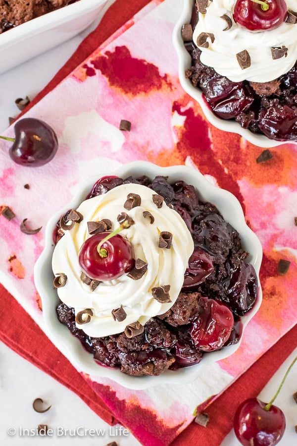 Overhead picture of a white bowl on a pink and orange towel filled with chocolate cherry cobbler and homemade whipped cream