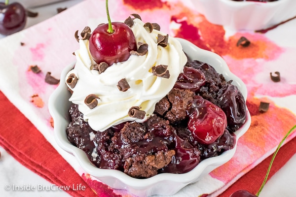 A white bowl on a pink and orange towel filled with chocolate cherry cobbler and whipped cream