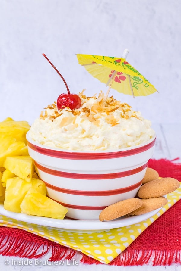 A white bowl with red striped filled with pina colada fruit dip and surrounded with pineapple wedges and vanilla wafers