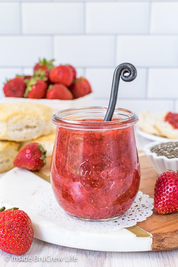A clear jar on a marble tray filled with strawberry chia jam and berries and biscuits behind it