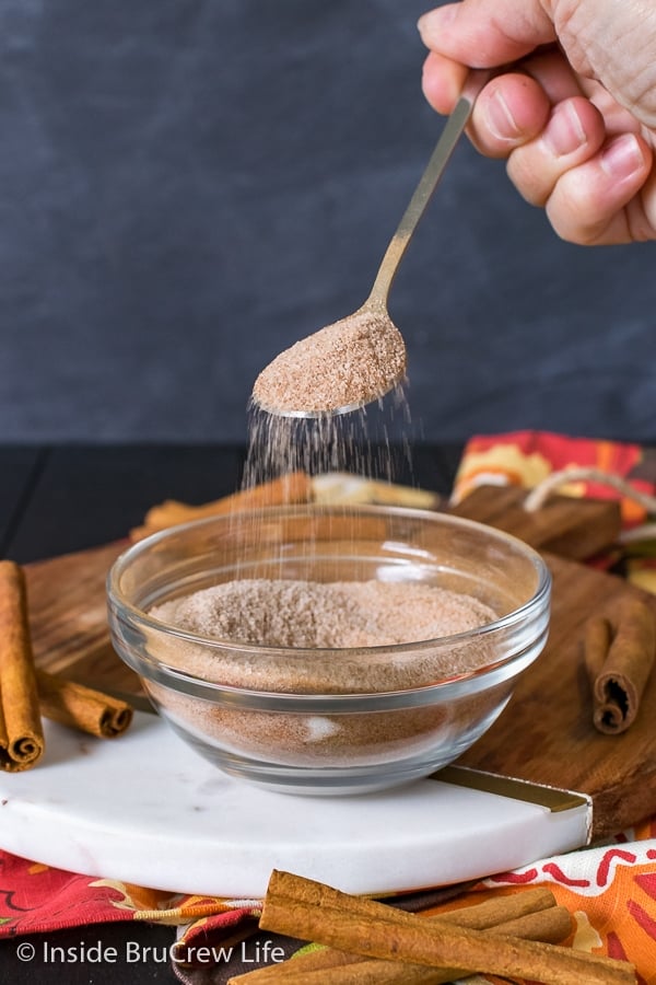 A clear bowl filled with cinnamon sugar with a spoon lifting sugar and and sprinkling it.