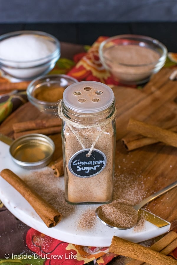 A clear jar with a shaker lid filled with cinnamon sugar sitting on a wood tray.