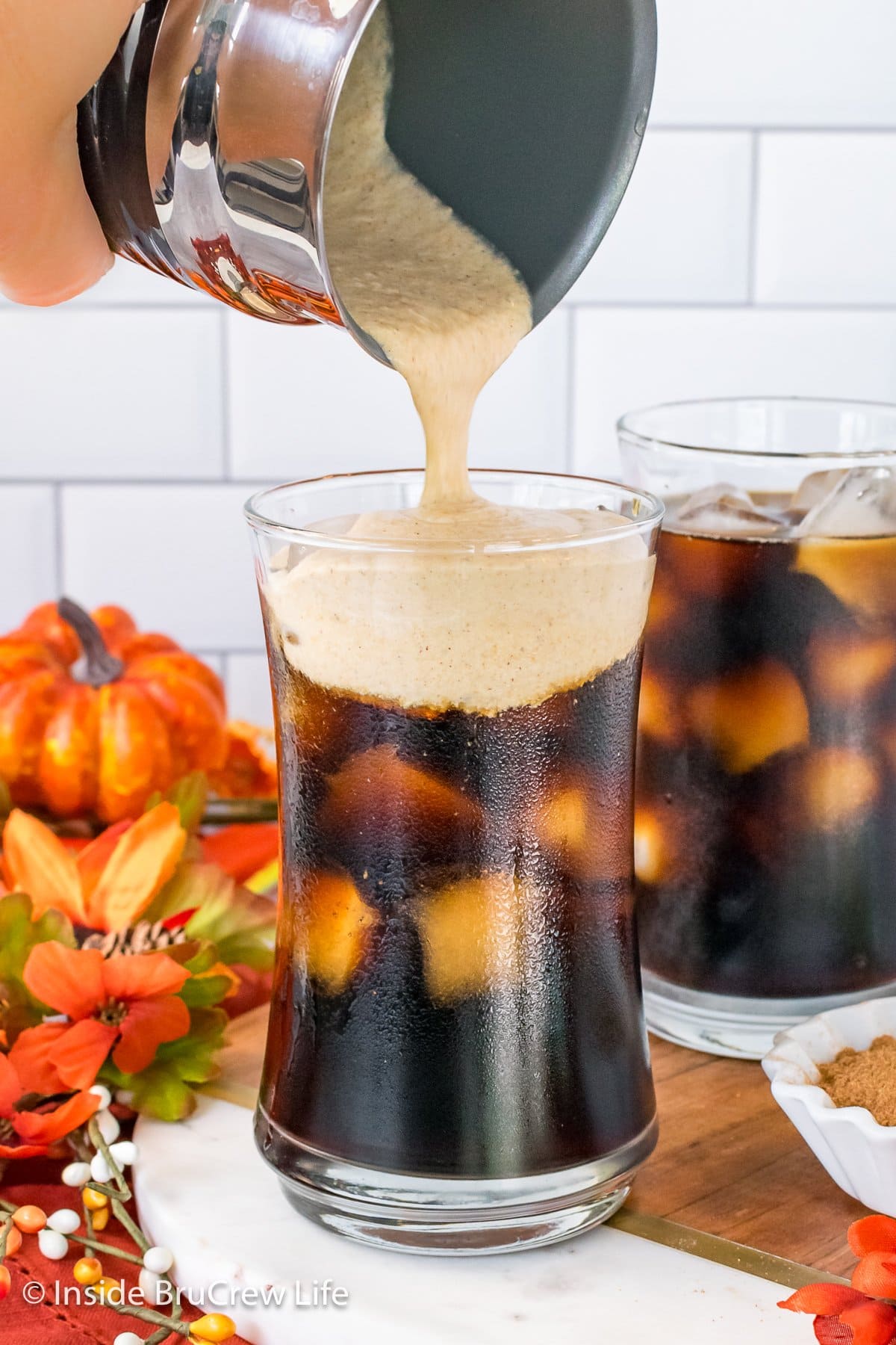 Two cups of iced coffee with cold foam being poured on top.