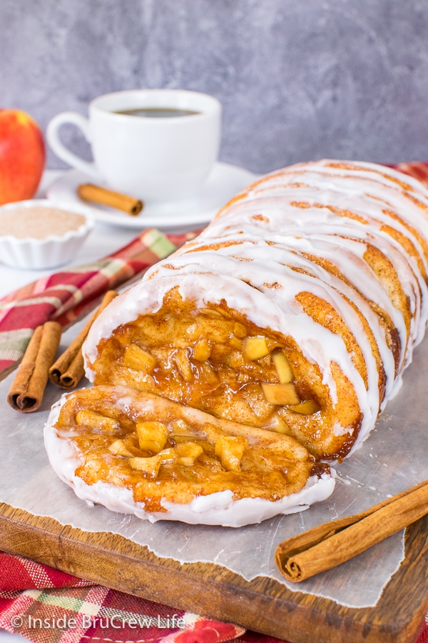 A wood board with a loaf of apple cinnamon pull apart bread drizzled with glaze and one sliced falling forward