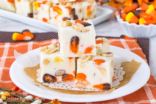 A white plate on an orange towel with three squares of candy corn fudge stacked on it and more fudge behind it