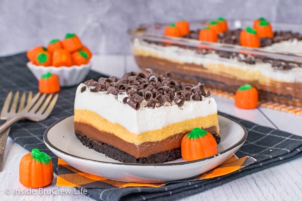 A square of chocolate pumpkin lasagna with chocolate curls on a white plate and the pan of dessert lasagna behind it