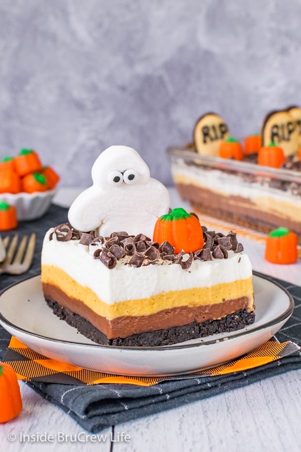 A white plate with a square of chocolate pumpkin lasagna topped with a marshmallow ghost and candy pumpkin