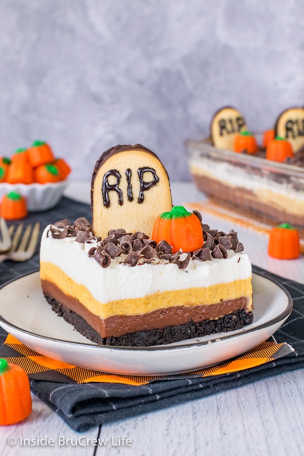 A white plate with a square of chocolate pumpkin lasagna topped with a tombstone cookie and candy pumpkin