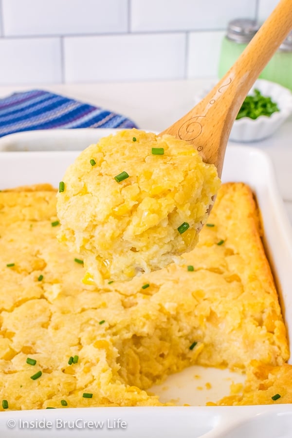 A white casserole dish with corn casserole in it and a wooden spoon lifting a scoop out of the pan.