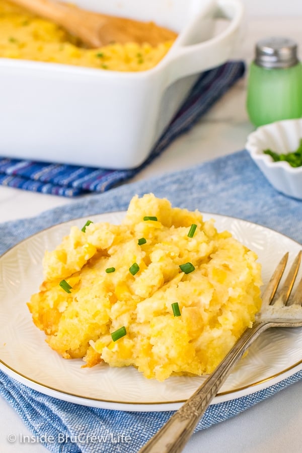 A white plate with scoop of Jiffy Corn Casserole on it with a fork and a white dish of corn casserole behind it.