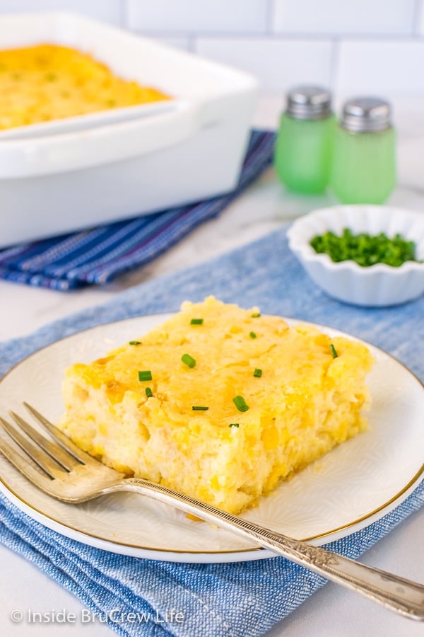 A white plate with a square of corn casserole and a fork on it and a white dish behind it.