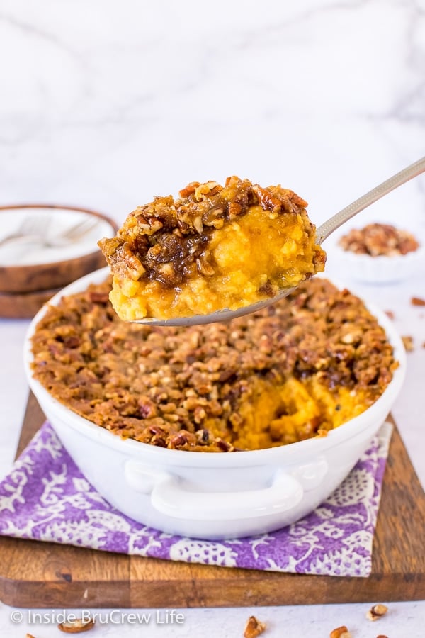 A white casserole dish on a purple towel with a spoon lifting out a scoop of sweet potato souffle