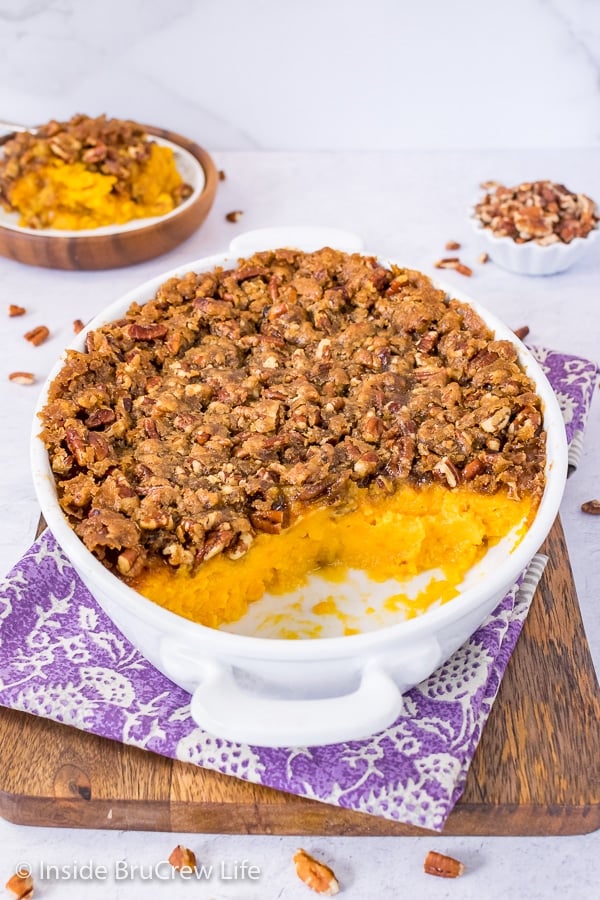 A white casserole dish on a purple towel filled with sweet potato souffle missing spoonfuls of casserole
