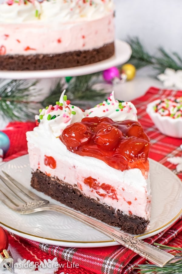 A white plate with a fork and slice of cherry mousse brownie cake on it and a cake plate with more cake behind it.