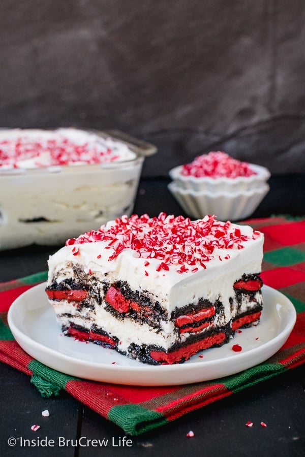 A white plate on a red and green towel with a piece of peppermint oreo icebox cake on it and a bowl of peppermint candies behind it