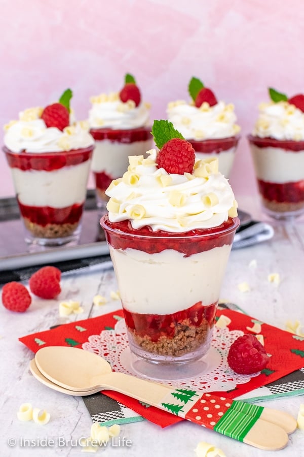 Five raspberry white chocolate cheesecake parfaits in clear cups on a white and pink background