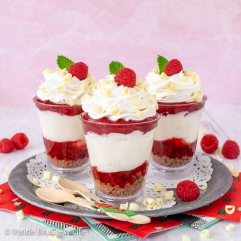 Three raspberry white chocolate cheesecake parfaits on a metal tray topped with whipped cream and a raspberry.