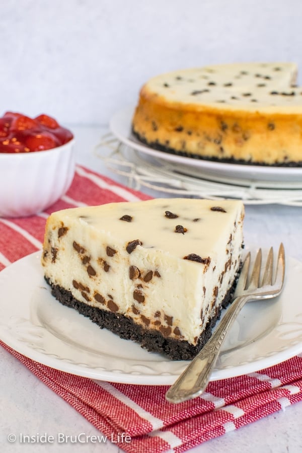 A slice of chocolate chip cheesecake on a white plate and more cheesecake behind it