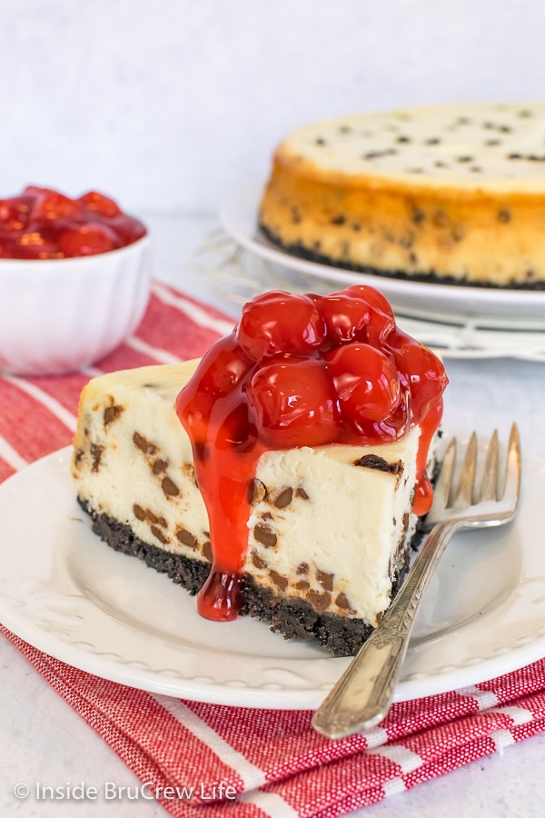A white plate with as slice of chocolate chip cheesecake topped with cherry pie filling on it