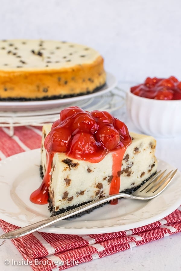A white plate with a slice of chocolate chip cheesecake with cherry pie filling on it and the full cheesecake behind it