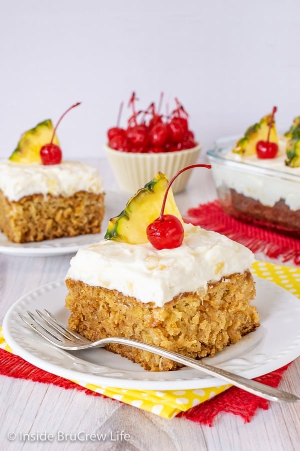 Two white plates with pineapple cake topped with pineapple cream cheese frosting, cherries, and pineapple slices.