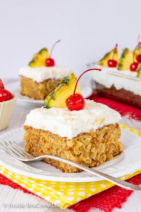 Two white plates with squares of pineapple sunshine cake topped with pineapple frosting and cherries on them